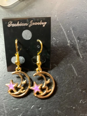 Guldörhängen med lila stjärna och måne - Ett par snygga örhängen i guldfärgad metall med hängande måne och en lila stjärna. Örhängena har en öppen design och krokar för enkel användning. Perfekta för dig som gillar unika detaljer och vill sticka ut lite extra.