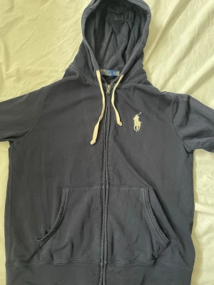 Mörkblå hoodie från Polo Ralph Lauren - Mörkblå hoodie med dragkedja från Polo Ralph Lauren. Klassisk design med vit broderad logga på bröstet, vita snören i huvan och två stora fickor framtill. Tillverkad i mjukt bomullsmaterial, perfekt för en avslappnad stil.