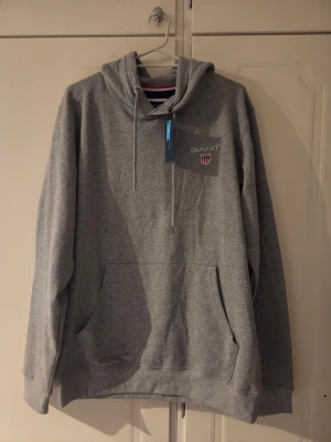 Grå hoodie från GANT med logga - Snygg grå hoodie från GANT med klassisk logga och sköld på bröstet. Hoodien har huva med dragsko, stor magficka och ribbade muddar. Tillverkad i mjukt sweatshirtmaterial som är perfekt för chill dagar. Enkel och stilren design som funkar till det mesta. Helt ny med taggs kvar.