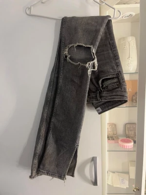 Grå slitna jeans från Zara - Säljer ett par grå jeans från Zara med slitningar och hål vid knäna. Jeansen har rak passform, klassiska fem fickor och råa benslut. Perfekta för en trendig och avslappnad stil.