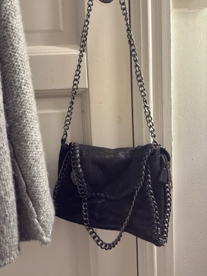 Svart axelväska med kedjedetaljer - Snygg svart axelväska med grova silverfärgade kedjor som axelrem och runt kanterna. Väskan har en vikbar lockdesign och är tillverkad i ett läderliknande material. Perfekt för dig som gillar en edgy och trendig stil.
