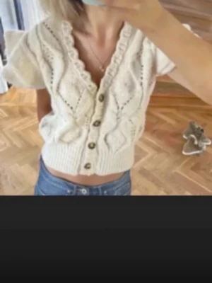 Vit stickad kortärmad kofta - Supersöt vit stickad kofta med korta puffärmar och djup v-ringning. Koftan har dekorativa hålmönster, volangkant längs ringningen och knäpps med fem bruna knappar framtill. Perfekt att styla med jeans eller kjol för en trendig look.