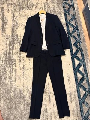 Mörkblå kostym med vit skjorta - Snygg mörkblå kostym med matchande byxor och kavaj samt en klassisk vit skjorta. Kavajen har två knappar och fickor framtill. Perfekt för dig som vill ha en stilren och tidlös look. Storlek är  140    9-12 år  priset går att diskutera 