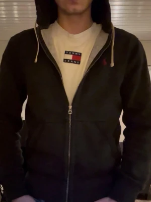 Svart hoodie från Polo Ralph Lauren - Svart hoodie med dragkedja från Polo Ralph Lauren. Klassisk design med huva och vita dragsnören, samt broderad logga på bröstet. Mjuk insida och ribbade muddar. Perfekt för en chill och stilren look.