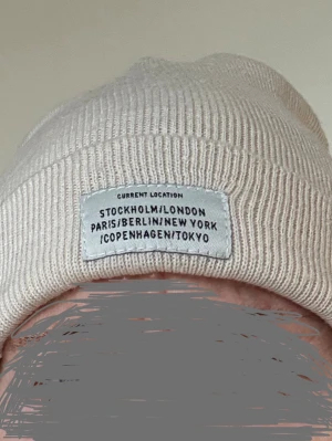 Ljusbeige mössa Gina onesize  - En stickad mössa ljusbeige. Mössan har en uppvikt kant och en rektangulär etikett med texten "CURRENT LOCATION STOCKHOLM/LONDON PARIS/BERLIN/NEW YORK /COPENHAGEN/TOKYO" fastsydd på framsidan. Gina tricot. Onesize. Använd ett fåtal ggr, är i ok skick, inga defekter, men spår av noppor. Polylana poly/ Akryl.