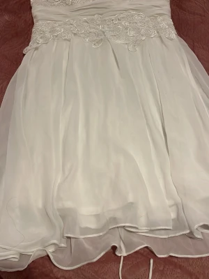 Vit kort klänning med spetsdetaljer - Säljer en vit kort klänning med vacker spets och snörning i midjan. Klänningen har ett lager av tunt tyg som ger en luftig känsla och fina broderade detaljer upptill. Perfekt för sommarens alla tillfällen eller studenten 