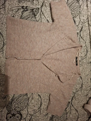 Beige kofta från H&M med pärlemorknappar - Mysig beige kofta från H&M med pärlemorskimrande knappar framtill. Koftan har en mjuk, stickad känsla och ribbade muddar vid ärmslut och nederkant. Perfekt att slänga över ett linne eller t-shirt för en avslappnad look.