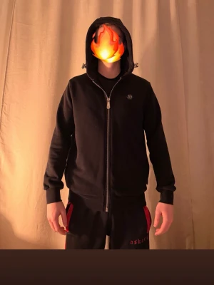 Svart hoodie från Philipp Plein - Snygg svart hoodie från Philipp Plein med dragkedja framtill och huva med dragsko. Har en diskret logga på bröstet och ribbade muddar vid ärmar och midja. Perfekt för en clean och stilren look. Passar M och L