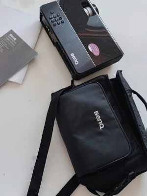 BenQ projektor med väska - Säljer en svart BenQ projektor med tillhörande bärväska och manual. Projektorn har en stilren, rektangulär design med knappar på ovansidan och tydlig BenQ-logga. Perfekt för filmkvällar eller presentationer. Väskan är svart med dragkedja och axelrem.