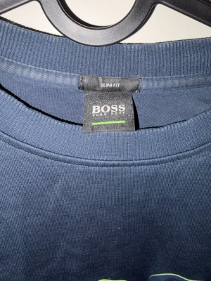 Blå sweatshirt från Hugo Boss - Mörkblå sweatshirt från Hugo Boss med rund halsringning och stort BOSS-tryck framtill. Slim fit-modell med ribbade muddar vid ärmslut och nederkant. Perfekt för en stilren och avslappnad look.