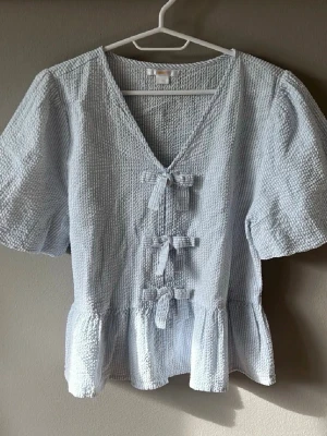 Ljusblå blus med rosetter - Supersöt ljusblå blus med struktur i tyget och tre dekorativa rosetter framtill. Blusen har v-ringning, korta puffärmar och volang nertill. 💕 från Gina Young! Storlek 170, använd en gång!