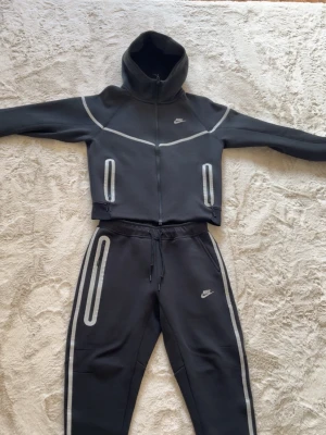 Nike tech fleece reflective  - Nike loungewear-set i svart med grå reflexdetaljer på både hoodie och byxor. Setet har dragkedja på tröjan, huva och två fickor framtill. Byxorna har dragsko i midjan och reflexränder längs benen. Perfekt för chill eller när du vill synas i mörkret. Vi kan diskutera priset.
