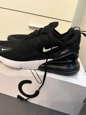 Nike Air Max 270 svarta sneakers - Svarta Nike Air Max 270 sneakers med vit sula och stor synlig Air-enhet i hälen. Ovandelen är i mesh och textil för bra andningsförmåga. Swoosh-logga i vitt på sidan och tungan. Snygg och sportig design som passar streetwear.