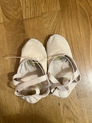 Ljusrosa balettskor med resårband - Säljer ett par ljusrosa balettskor med resårband över foten och delad mockasula undertill. Skorna är mjuka och flexibla, perfekta för dans eller balett. Klassisk design med rund tå och elastisk kant för bra passform.