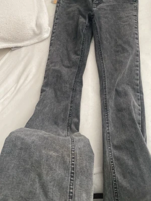 Grå bootcut jeans från Gina Tricot - Snygga grå bootcut jeans från Gina Tricot med tvättad look och klassiska fem fickor. Jeansen har hög midja och utsvängda ben, perfekta för en trendig och avslappnad stil. Materialet är mjukt denim med stretch för extra komfort.