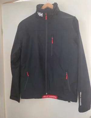 Svart jacka från Helly Hansen - Svart vindjacka från Helly Hansen med röda dragkedjor och logga på krage.  Strl S 