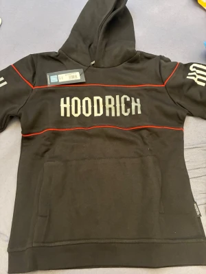 Svart HOODRICH hoodie med röda detaljer - Svart hoodie från Hoodrich med stora vita tryck på bröstet och ärmen. Hoodien har röda linjer som detaljer och en klassisk huva. Stor magficka framtill och mjukt material som känns skönt mot huden. Perfekt för en streetwear-look. Den är helt oanvänd, prislapp finns kvar. 