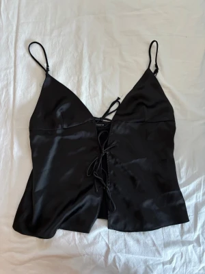 Svart satinlinnetopp från Zara - Svart linnetopp från Zara i glansigt satinmaterial med smala axelband och snygg snörning framtill. Toppen har en croppad passform och djup v-ringning, perfekt för att lyfta vilken outfit som helst.