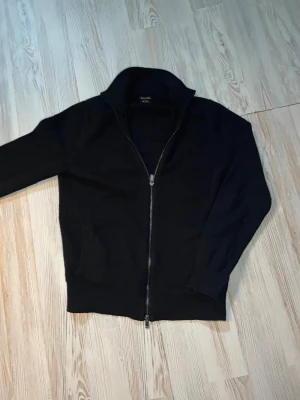 Svart ziptröja från Massimo Dutti - Stilren svart ziptröja från Massimo Dutti med hög krage och hel dragkedja. Tröjan är stickad och har ribbade muddar vid ärmslut och nederkant. Perfekt för lager-på-lager och enkel att matcha med olika outfits