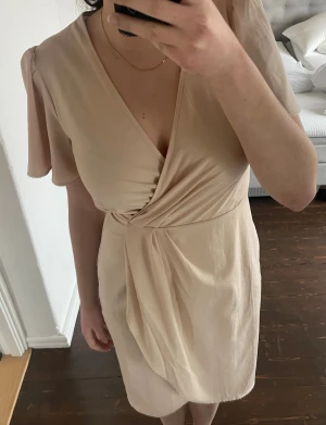 Beige omlottklänning från H&M - Snygg beige omlottklänning från H&M med v-ringning och korta, fladdriga ärmar. Klänningen har en draperad effekt framtill och är tillverkad i ett mjukt, lätt tyg som känns skönt mot huden. Perfekt för sommarens alla tillfällen.