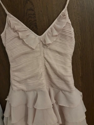 Ljusrosa volangklänning  - Söt ljusrosa kortklänning med smala axelband och volangdetaljer både upptill och nertill. Klänningen har en v-ringad urringning och är sydd i ett lätt, plisserat tyg som ger en romantisk känsla. Perfekt för sommarens alla tillfällen.