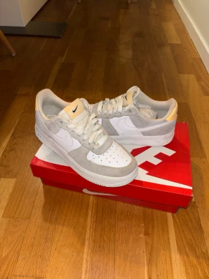 Nike Air Force 1 vita/grå sneakers - Nike Air Force 1 sneakers i vitt och ljusgrått med beige detaljer. Klassisk låg modell med ovandel i läder och mocka, perforerad tå och vit sula. Swoosh-logga på sidan och Nike-märke på hälen. Perfekta för en clean och stilren look.