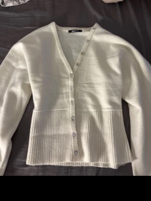 Vit ribbad kofta från Gina Tricot - Stilren vit kofta från Gina Tricot med v-ringning och knappar framtill. Koftan har långa ärmar och en bred ribbad kant nedtill som ger en snygg kontrast. Perfekt att slänga över en topp för en enkel men chic look. Använd 1 gång!