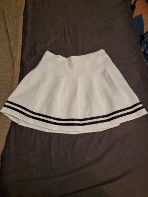 Vit kortkjol med svarta ränder SHEIN - Vit kortkjol från SHEIN med två svarta ränder längs nederkanten. Kjolen har en klassisk tennisstil och är lätt utställd för en snygg siluett. Perfekt för dig som gillar sportig och trendig look. För barn men passar XS-S