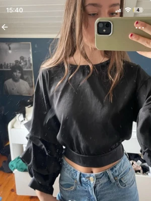 Svart croppad topp med volangärmar - Säljer en svart croppad topp med bred resår i midjan och volangdetaljer på ärmarna. Toppen har rund hals och långa ärmar, perfekt för att skapa en trendig look. Passar dig som gillar att sticka ut med coola detaljer.