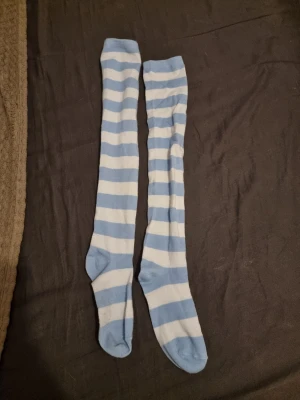 Blåvita randiga overknee-strumpor - Säljer ett par långa overknee-strumpor med breda blå och vita ränder. Strumporna är mjuka och stretchiga, perfekta för att ge en lekfull touch till din outfit. Materialet känns bekvämt mot huden och de sitter uppe bra på benet.