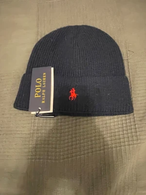 Marinblå stickad mössa Polo Ralph Lauren - Snygg marinblå stickad mössa från Polo Ralph Lauren med klassisk röd logga broderad framtill. Perfekt för kalla dagar och enkel att matcha med olika outfits. Tidlös design och mjukt material.