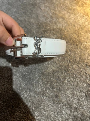 Vitt YSL bälte i skinn med silverdetaljer - Stilrent vitt bälte från Yves Saint Laurent i skinn med klassiskt YSL-logo i silver framtill. Bältet har en silverfärgad spänne och är perfekt för att lyfta vilken outfit som helst med en lyxig touch.