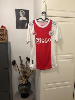 Ajax Amsterdam matchtröja Adidas S - Ajax Amsterdam matchtröja från Adidas i storlek S. Tröjan har klassisk röd mittpanel och vita sidor, med Ajax-logga och Ziggo sponsortryck på bröstet. Tillverkad i Primegreen polyester med autentisk licensmärkning. Perfekt för fotbollsfans!