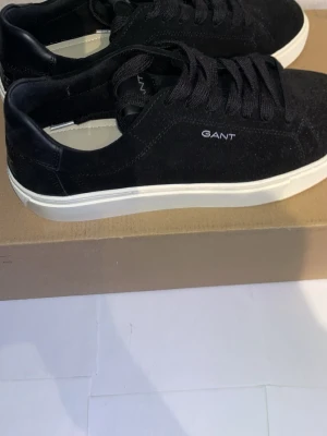 Svarta sneakers från Gant i mocka - Stilrena svarta sneakers från Gant med ovandel i mjuk mocka och vita sulor. Klassisk låg modell med svarta skosnören och diskret Gant-logga på sidan och plösen. Insidan är ljus och ger en snygg kontrast till det svarta yttermaterialet.