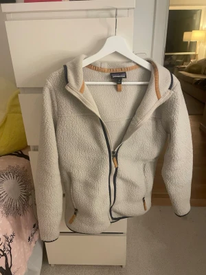 Vit teddyjacka från Patagonia - Mysig vit teddyjacka från Patagonia med hel dragkedja och två fickor framtill. Jackan har kontrastdetaljer i beige och mörkblått vid dragkedjor och krage. Tillverkad i fluffigt teddy-material som håller dig varm och bekväm. Perfekt för kyliga dagar.