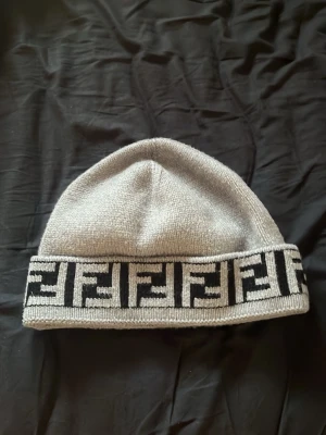 Grå Fendi mössa med logotypkant - Snygg grå stickad mössa från Fendi med bred uppvikt kant där Fendis ikoniska logotypmönster syns i svart och grått. Klassisk form och mjukt material som håller dig varm. Perfekt accessoar för att lyfta din streetstyle.