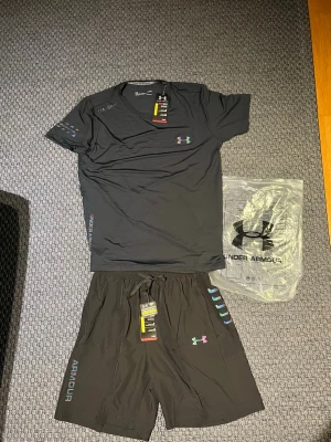 Under armour träningsset  - Snyggt svart träningsset från Under Armour med t-shirt och shorts. Perfekt som träningskläder men också till sommaren. Storleken är L men den passar perfekt som M. Ifall ni har några funderingar så är det bara att fråga.