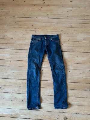 Blå raka jeans från Nudie Jeans - Säljer ett par klassiska blå jeans från Nudie Jeans med rak passform och fyra fickor. Jeansen har kontrastsömmar och patch i brunt läder bak i midjan. Tillverkade i slitstark denim, perfekta till vardagslooken. Riktigt bra skick, omöjligt att se att de har använts. Ny pris c:a 1400kr