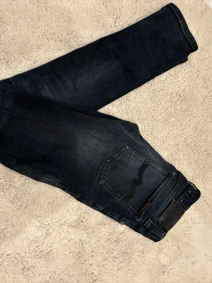 Mörkblå skinny jeans från Nudie - Säljer ett par mörkblå skinny jeans från Nudie med klassisk femficksdesign och svarta sömmar på bakfickan. Jeansen har en smal passform och är tillverkade i stretchigt denimtyg för extra komfort.