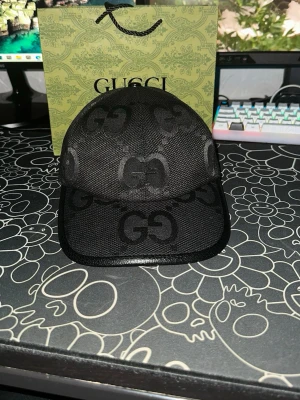 Svart Gucci keps  - Säljer en svart keps från Gucci med det klassiska GG-mönstret över hela kepsen. Kepsen har böjd skärm och justerbart band bak. Tillverkad i mesh-liknande material som ger en luftig känsla och modern look. Perfekt för dig som vill ha något stilrent och exklusivt.