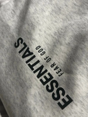 Grå shorts Essentials Fear of God - Säljer ett par ljusgrå shorts från Essentials Fear of God med svart logotyp på benet. Shorts med snörning i midjan och sidofickor, tillverkade i mjuk bomull. Perfekt för en avslappnad och trendig look. Är medium men sitter som small