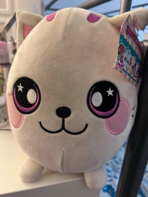 Squishmallows katt plyschleksak - Supermjuk och rund plyschleksak från Squishmallows som föreställer en katt. Den är vit med rosa detaljer på öron, kinder och ögon, samt har broderade svarta och rosa stjärnögon. Perfekt att krama eller ha som dekoration i rummet.