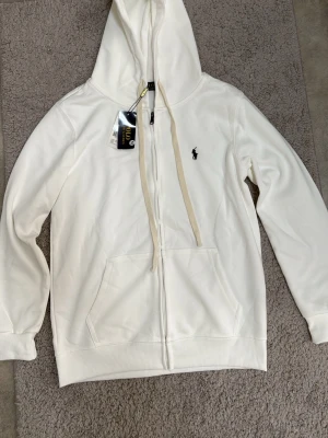 Vit hoodie från Polo Ralph Lauren - Snygg vit hoodie från Polo Ralph Lauren med dragkedja framtill, två stora fickor och klassisk huva med snören. Ikoniska lilla loggan broderad på bröstet. Tillverkad i mjukt material som känns skönt mot huden. Perfekt för en avslappnad och stilren look.