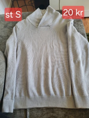 Ljusgrå stickad polotröja - Ljusgrå stickad polotröja med lång ärm och bred ribbad mudd vid ärmslut och nederkant. Tröjan har en hög, lös polokrage och är enkel i designen utan synliga loggor eller mönster. Perfekt för lager på lager och en clean look.