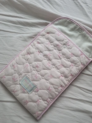 Quiltad datafodral i hjärtformer med rosettryck - Söt quiltad dator fodral i ljusrosa med hjärtformat mönster och små rosa rosetter.   insidan har två fack för datorn och eventuella föremål och en knapp att stänga med upptill. Perfekt för skolan!