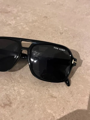 Svarta Tom Ford solglasögon - Snygga svarta solglasögon från Tom Ford med klassisk fyrkantig form och coola detaljer i guld på bågarna. Glasen är mörka och ramen är gjord i syntetmaterial. Perfekt för dig som vill ha en stilren och trendig look.
