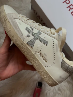 premiata shoes - Stevend 8127. snygga vita sneakers från Premiata med grå glitterdetaljer på sidan och hälen. Skorna har en låg siluett, beige mockadetaljer och text på sulan. Klassisk snörning och stilren design som passar till många outfits. aldrig buren