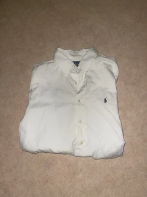 Vit skjorta från Ralph Lauren - Klassisk vit skjorta från Ralph Lauren med button-down krage och broderad logga på bröstet. Skjortan har lång ärm, knappar framtill och är tillverkad i bomull. Perfekt till en clean och stilren look.