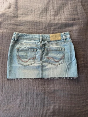 Ljusblå lågmidjad jeanskjol - Snygg ljusblå jeanskjol med rå, fransig kant nertill och klassiska fickor både fram och bak. Kjolen har bälteshällor och en liten broderad detalj på bakfickorna. Perfekt för en avslappnad och trendig look.
