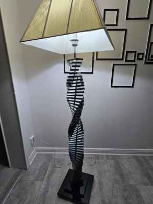 Modern golvlampa med spiralformad fot - Unik golvlampa med spiralformad fot i svart och silverfärgat material. Lampan har en fyrkantig fot och en beige lampskärm med trästruktur. Perfekt som statement piece i vardagsrummet eller sovrummet.
